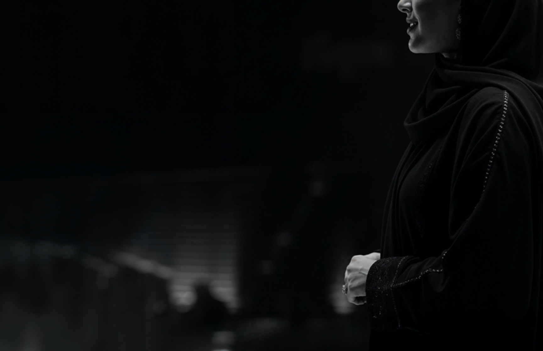 Side view of a woman in a black abaya speaking, with a dark blurred background